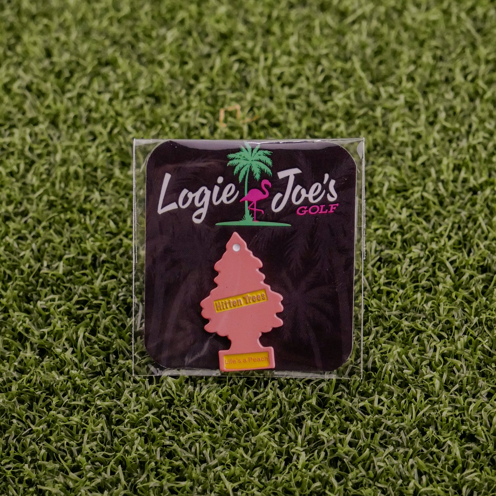 A pink tree-shaped air freshener labeled “Hitten Trees Life’s a Peach” by Logie Joe’s Golf sits on artificial green grass, bringing a playful vibe to funny golf accessories.