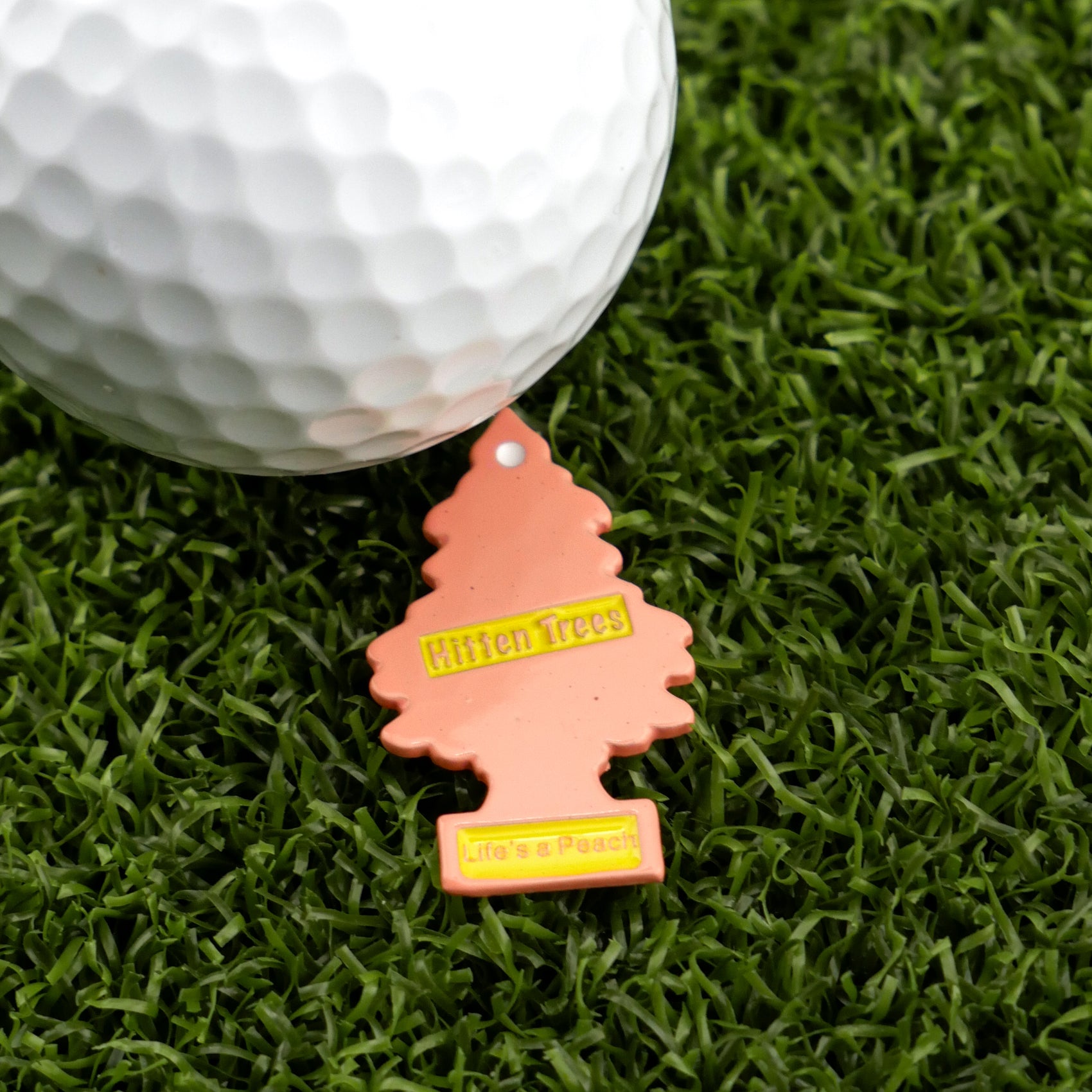 A white golf ball rests on green turf beside Logie Joe's Golf Hitten Trees Life’s a Peach, a peach-colored tree-shaped air freshener—ideal for fans of funny golf accessories and unique golf ball markers.