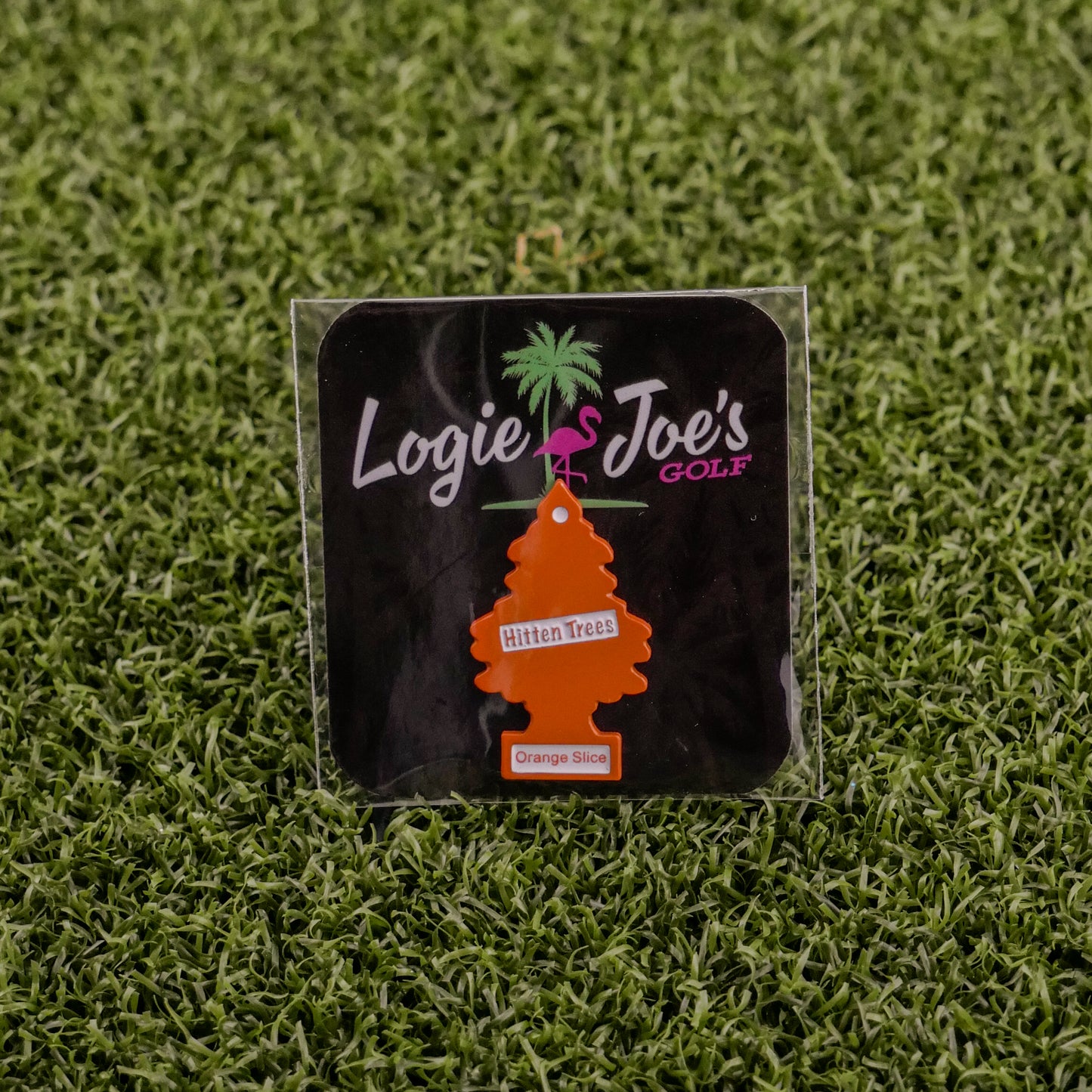 The Hitten Trees Orange Slice air freshener from Ballmarkers.com, shaped like an orange tree and featuring the Logie Joes Golf logo, is a funny golf accessory shown on artificial green grass.