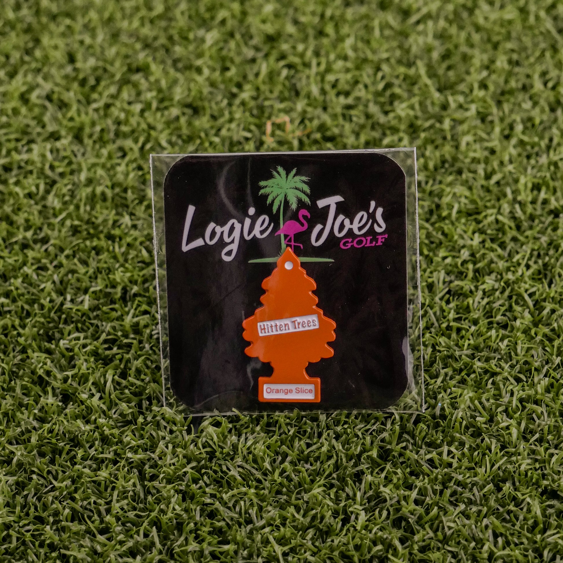 The Hitten Trees Orange Slice air freshener from Ballmarkers.com, shaped like an orange tree and featuring the Logie Joes Golf logo, is a funny golf accessory shown on artificial green grass.