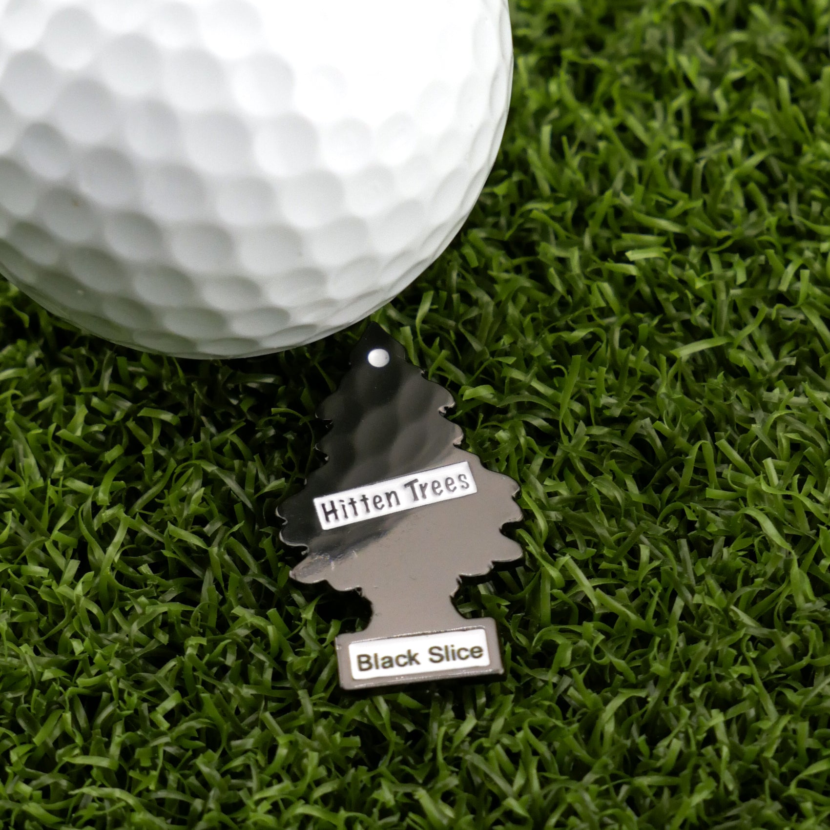 A white golf ball rests on green artificial grass beside a tree-shaped black tag labeled "Hitten Trees" and "Black Slice"—a fun accessory from Logie Joe's Golf to add personality to your game.