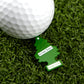 A white golf ball sits on artificial grass beside a green tree-shaped "Hitten Trees NutNBut Trees" keychain from Logie Joe's Golf—perfect for fans of humorous golf accessories.
