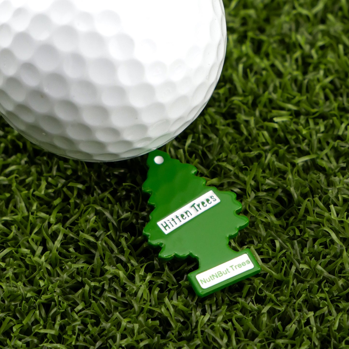 A white golf ball sits on artificial grass beside a green tree-shaped "Hitten Trees NutNBut Trees" keychain from Logie Joe's Golf—perfect for fans of humorous golf accessories.
