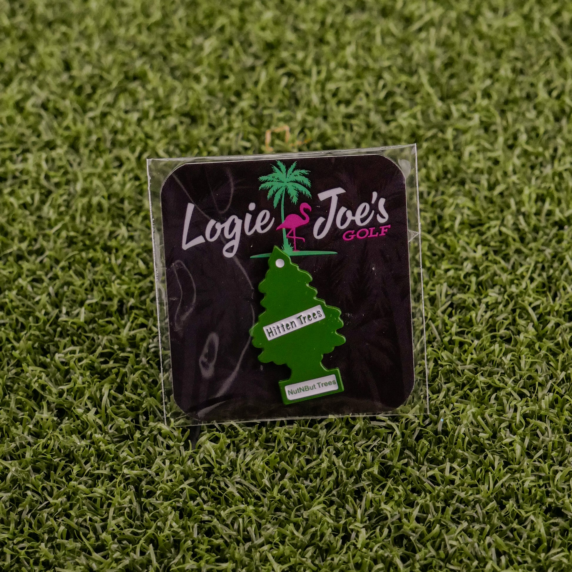 A green, tree-shaped ball marker labeled Hitten Trees NutNBut Trees by Ballmarkers from Logie Joes is attached to a black card with brand text—an amusing gift idea for golf lovers, displayed on artificial grass.