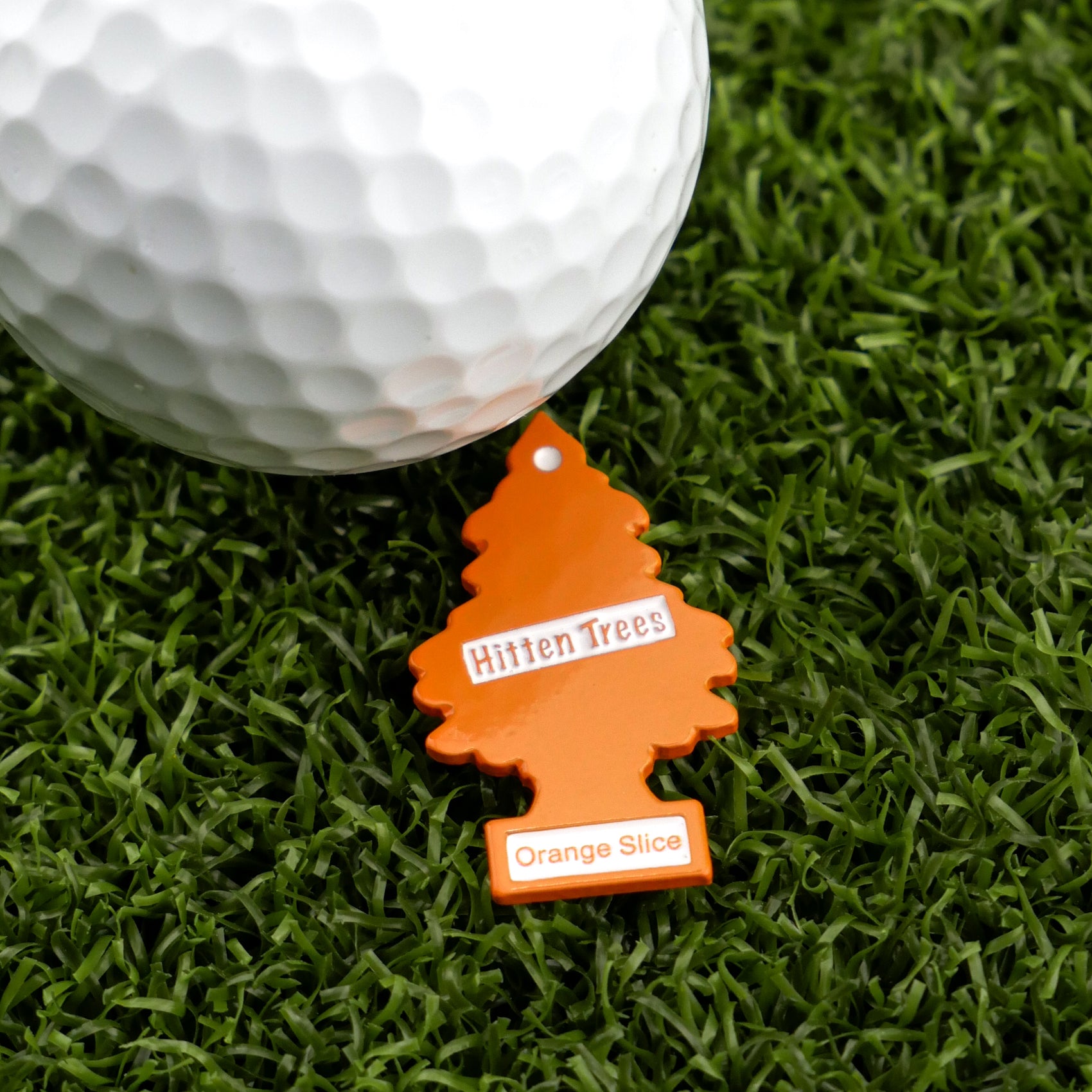 The Hitten Trees Orange Slice keychain from Ballmarkers.com, shaped like an orange tree and labeled with its name, rests on green turf beside a white golf ball—ideal for those seeking unique or humorous golf ball markers.