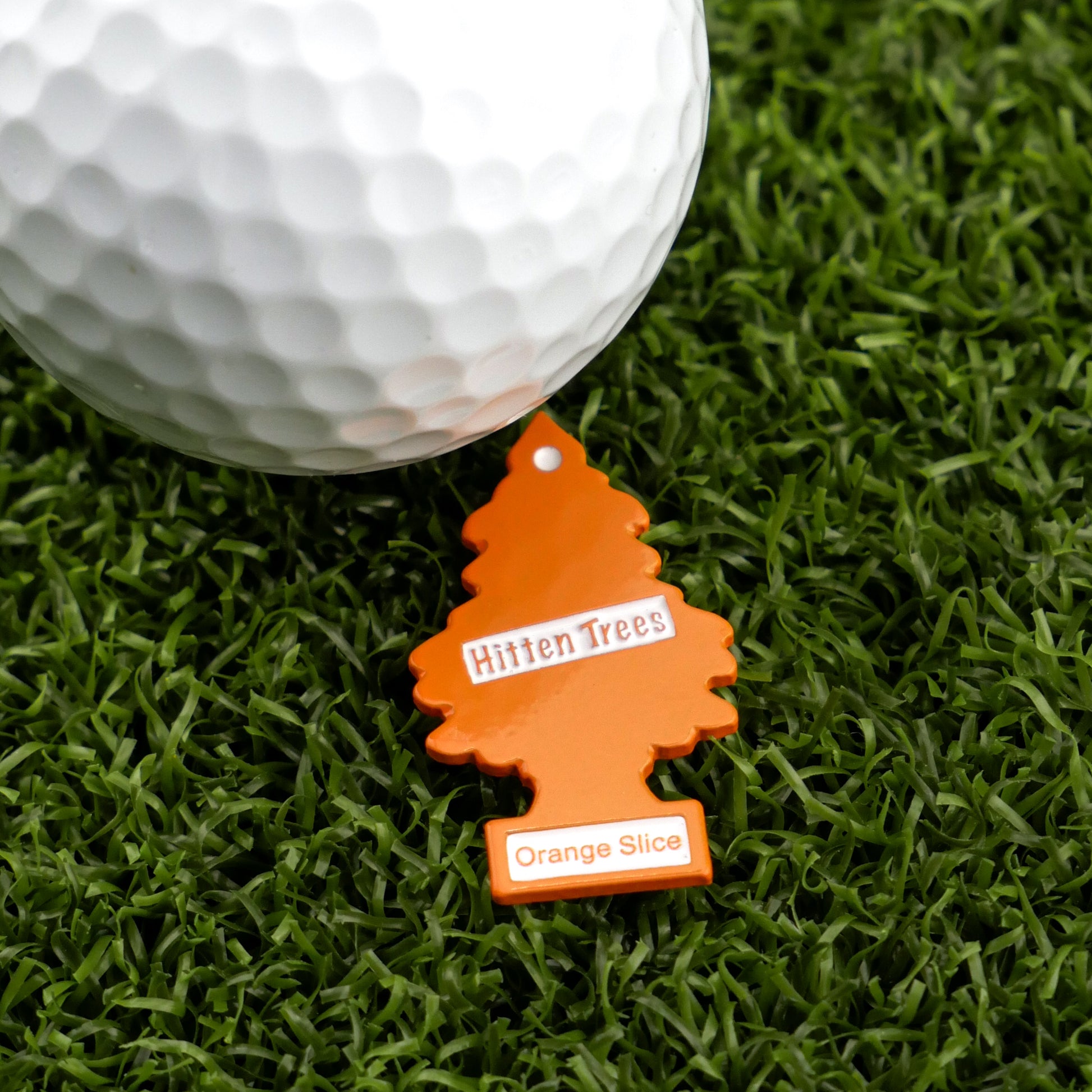 The Hitten Trees Orange Slice keychain from Ballmarkers.com, shaped like an orange tree and labeled with its name, rests on green turf beside a white golf ball—ideal for those seeking unique or humorous golf ball markers.