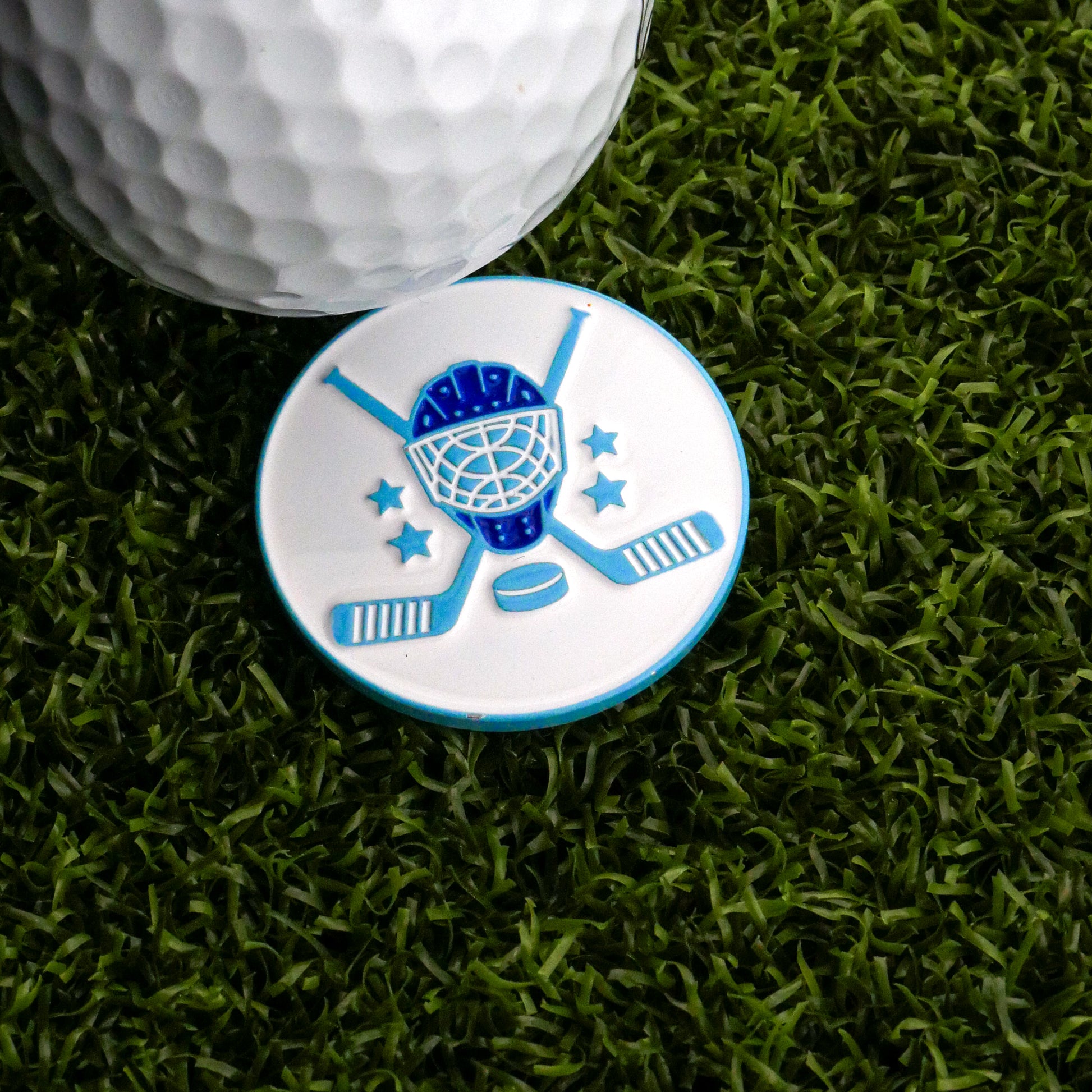 A white golf ball sits on artificial grass next to the Ballmarkers.com Hockey Equipment Ball Marker, featuring a blue goalie mask, crossed sticks, stars, and a puck—a tribute to classic hockey gear.
