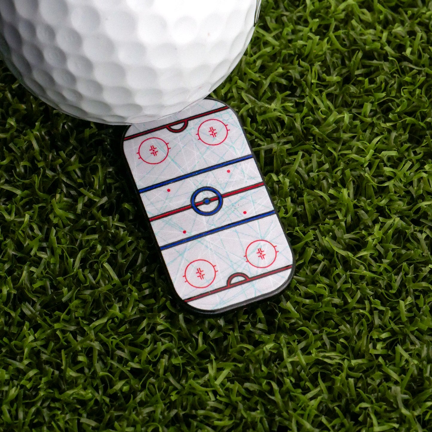 A golf ball sits on artificial grass next to the Ballmarkers.com Hockey Rink Ball Marker, which displays an ice hockey rink illustration and brings championship intensity to the green.