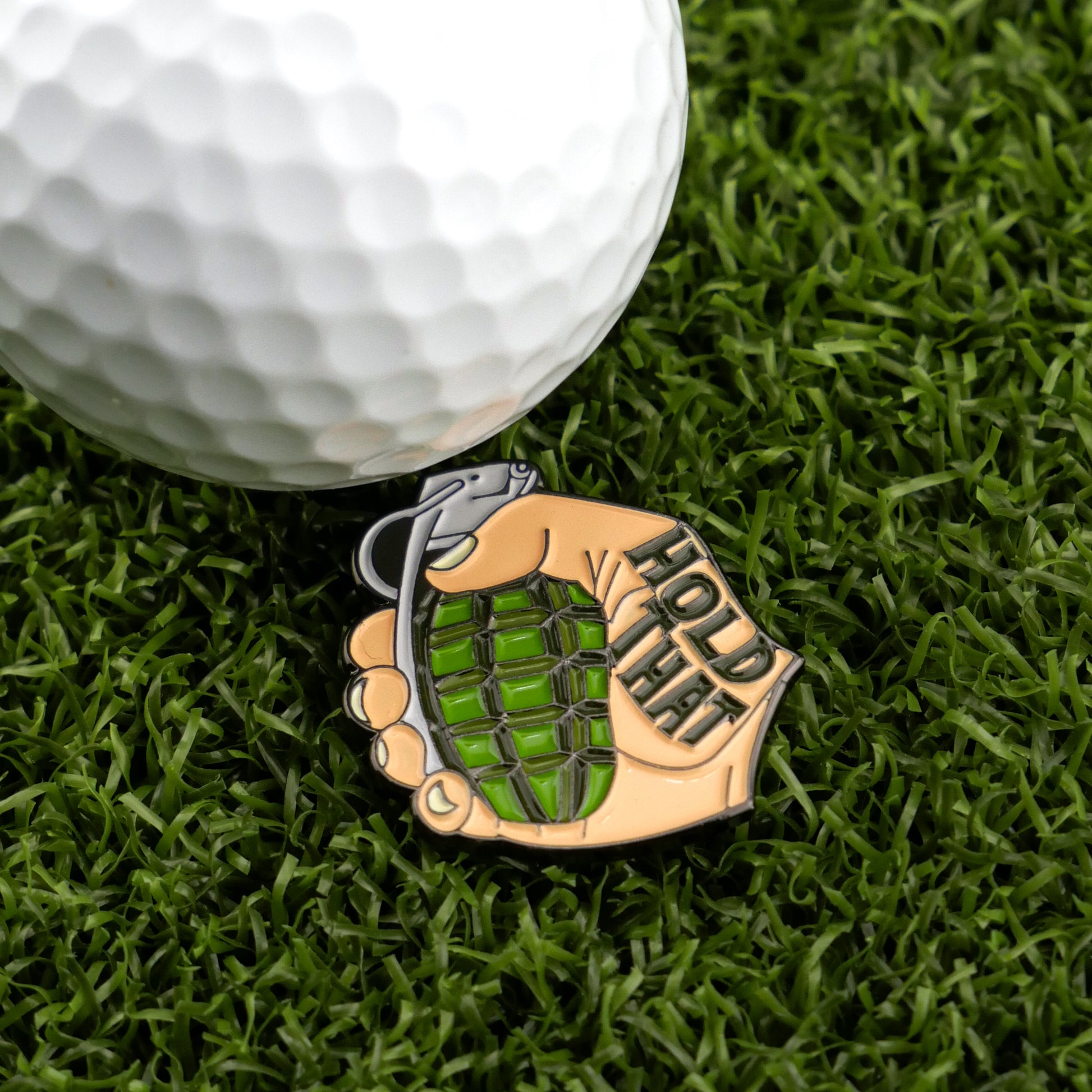 A close-up of a golf ball on artificial grass beside Logie Joe’s Golf Hold That Grenade—a unique hand-grenade shaped marker perfect for those who love standout golf accessories.