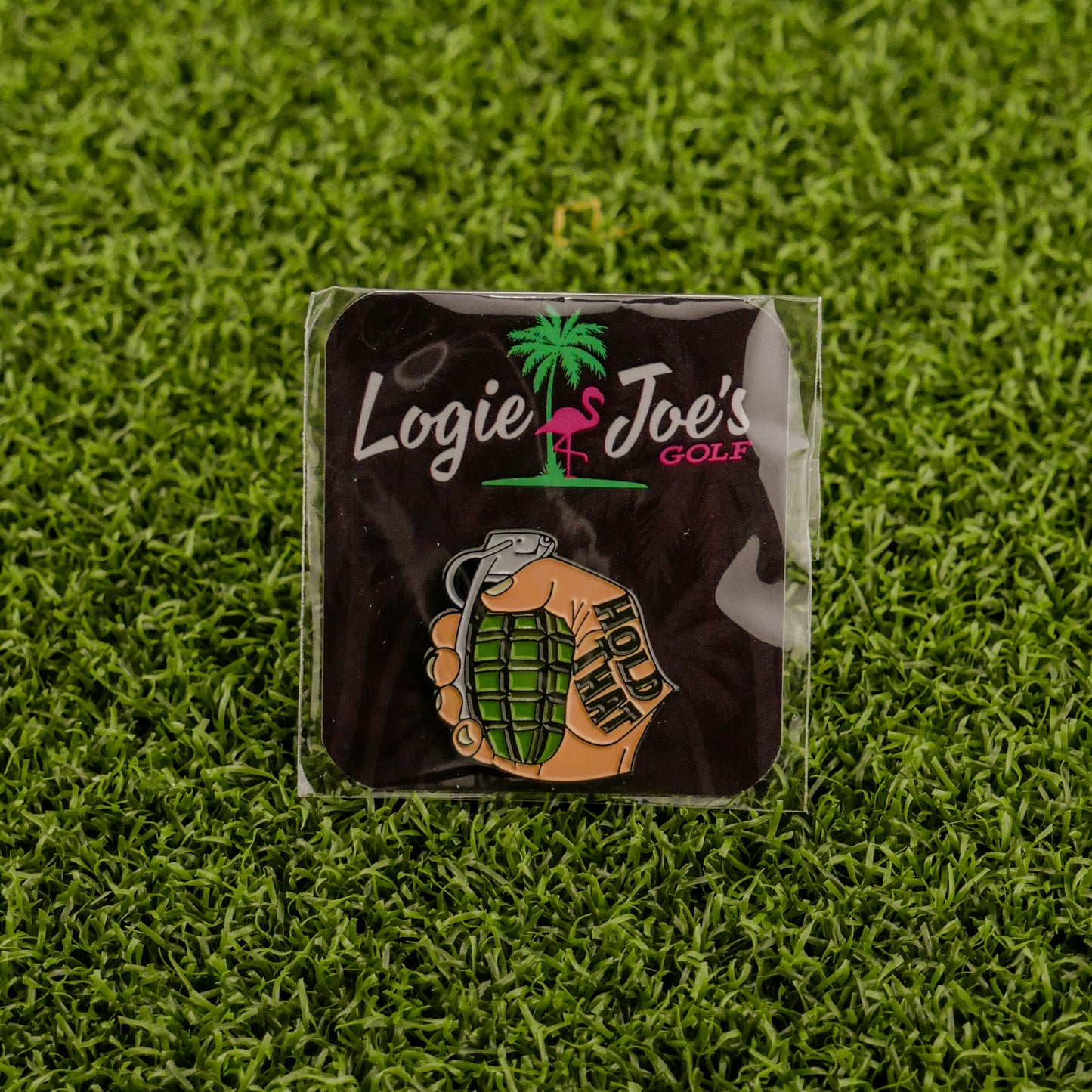 The "Hold That Grenade" pin by Logie Joe's Golf shows a hand with HOLD THIS gripping a grenade, set on artificial grass with a brand card—an edgy addition to any premium golf accessory or unique ball marker collection.