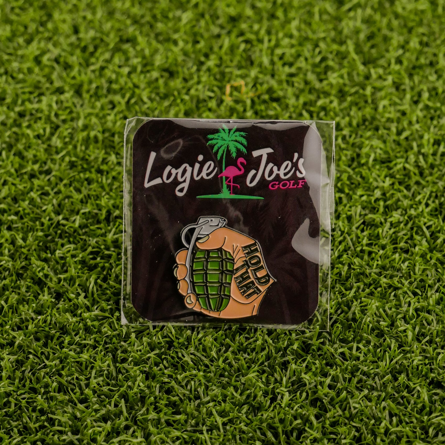 The "Hold That Grenade" pin by Logie Joe's Golf shows a hand with HOLD THIS gripping a grenade, set on artificial grass with a brand card—an edgy addition to any premium golf accessory or unique ball marker collection.