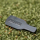 A small, black, bottle-shaped Louisiana Hot Sauce Ball Marker by Pins and Aces rests on green artificial turf.