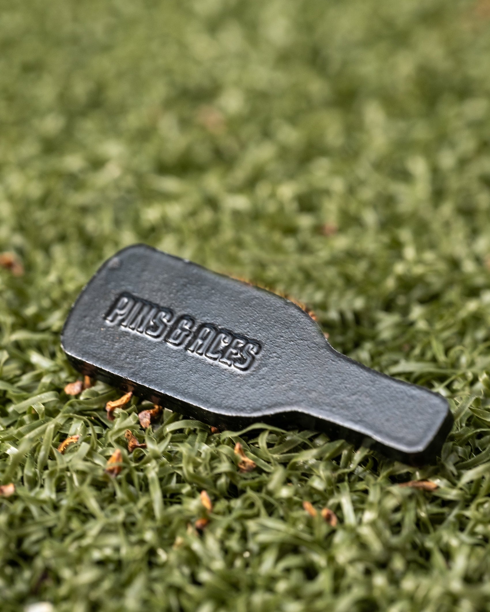 A small, black, bottle-shaped Louisiana Hot Sauce Ball Marker by Pins and Aces rests on green artificial turf.