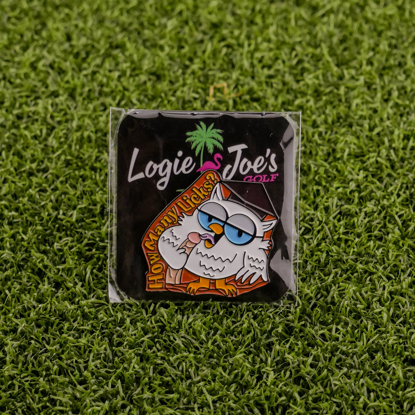 A square pin called "How many licks? Owl" from Ballmarkers.com features a cartoon owl with orange text on artificial grass. Packaged with Logies Joes Golf branding and a green palm tree, it’s ideal for fans of funny golf accessories.