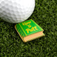 A white golf ball and Logie Joe's Golf "How to Putt" enamel pin shaped like a book rest on artificial grass, serving as a unique and charming golf ball marker for your setup.