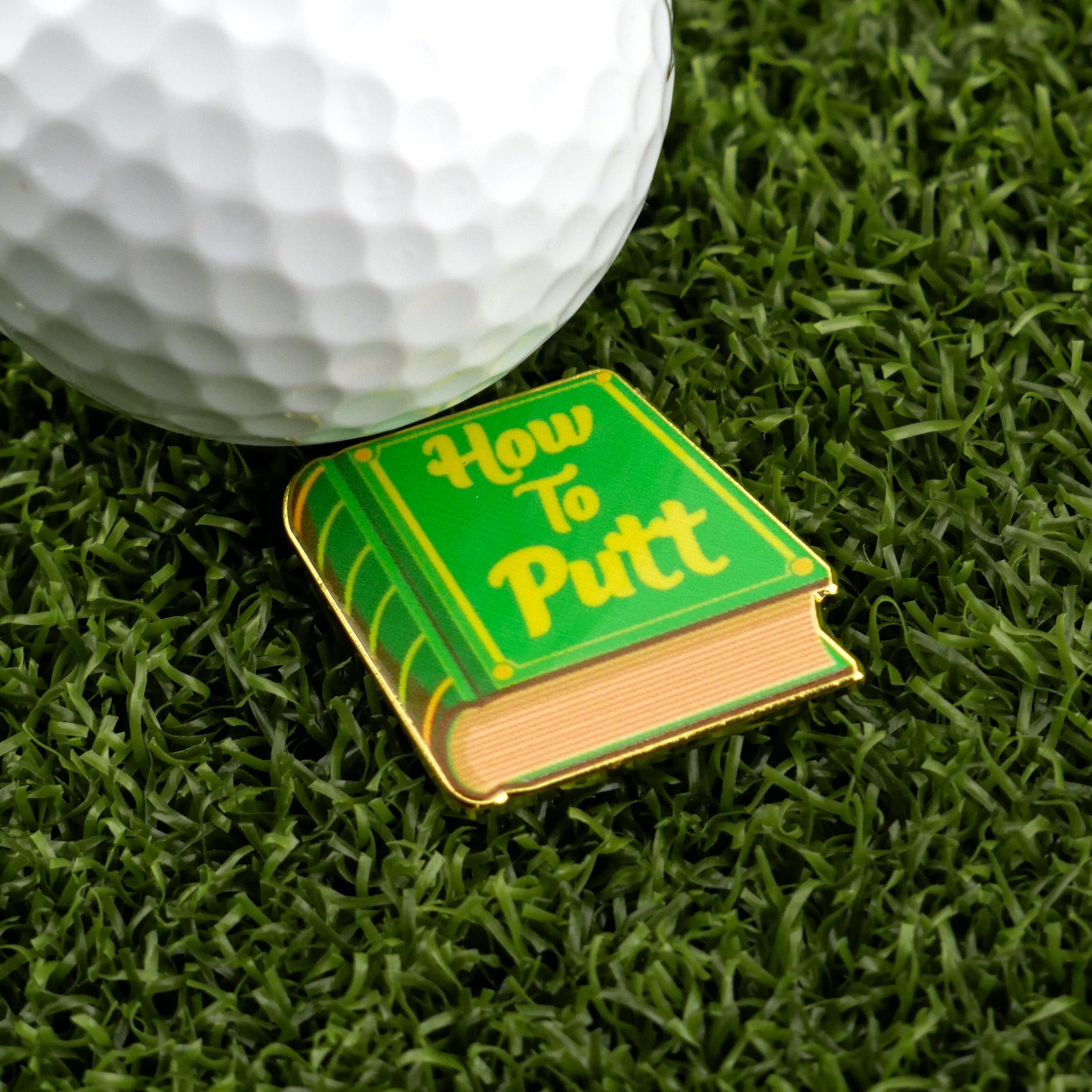 A white golf ball and Logie Joe's Golf "How to Putt" enamel pin shaped like a book rest on artificial grass, serving as a unique and charming golf ball marker for your setup.