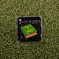 The How to Putt enamel pin by Logie Joe's Golf doubles as a premium ball marker. Shaped like a green book, it’s displayed on black Logie Joe's packaging atop artificial grass, making it a standout golf accessory.