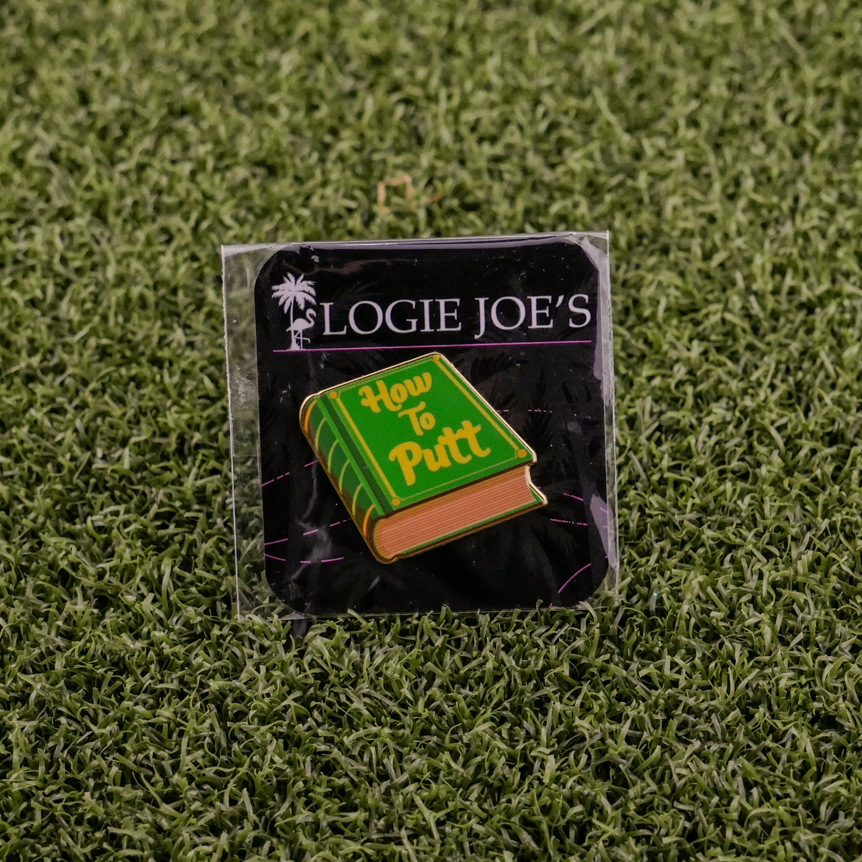 The How to Putt enamel pin by Logie Joe's Golf doubles as a premium ball marker. Shaped like a green book, it’s displayed on black Logie Joe's packaging atop artificial grass, making it a standout golf accessory.