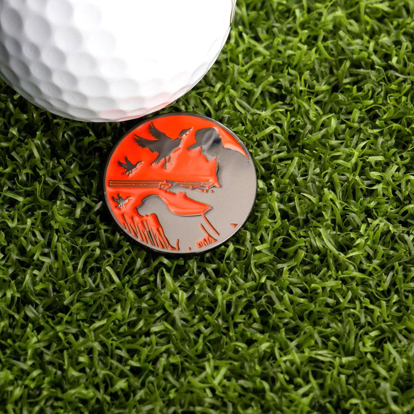 A close-up of Logie Joe's Golf Duck Hunter Ball Marker, a unique golf accessory with red and silver bird and nature designs, shown on green artificial grass—perfect for elevating your short game.