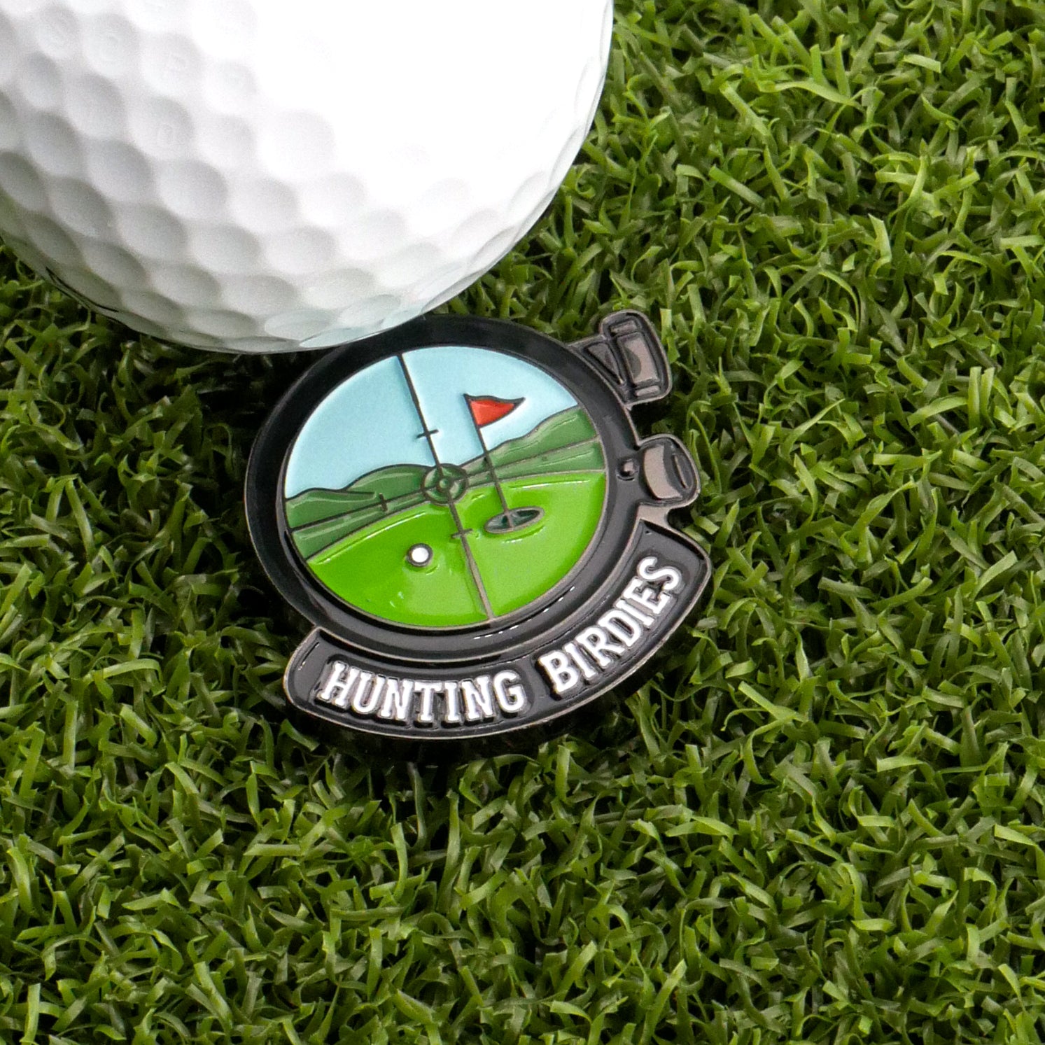 A golf ball sits on green grass beside the Logie Joe's Golf Hunting Birdies Ball Marker, which features a golf course illustration, a red flag, and the text HUNTING BIRDIES—a top funny golf accessory for your game.