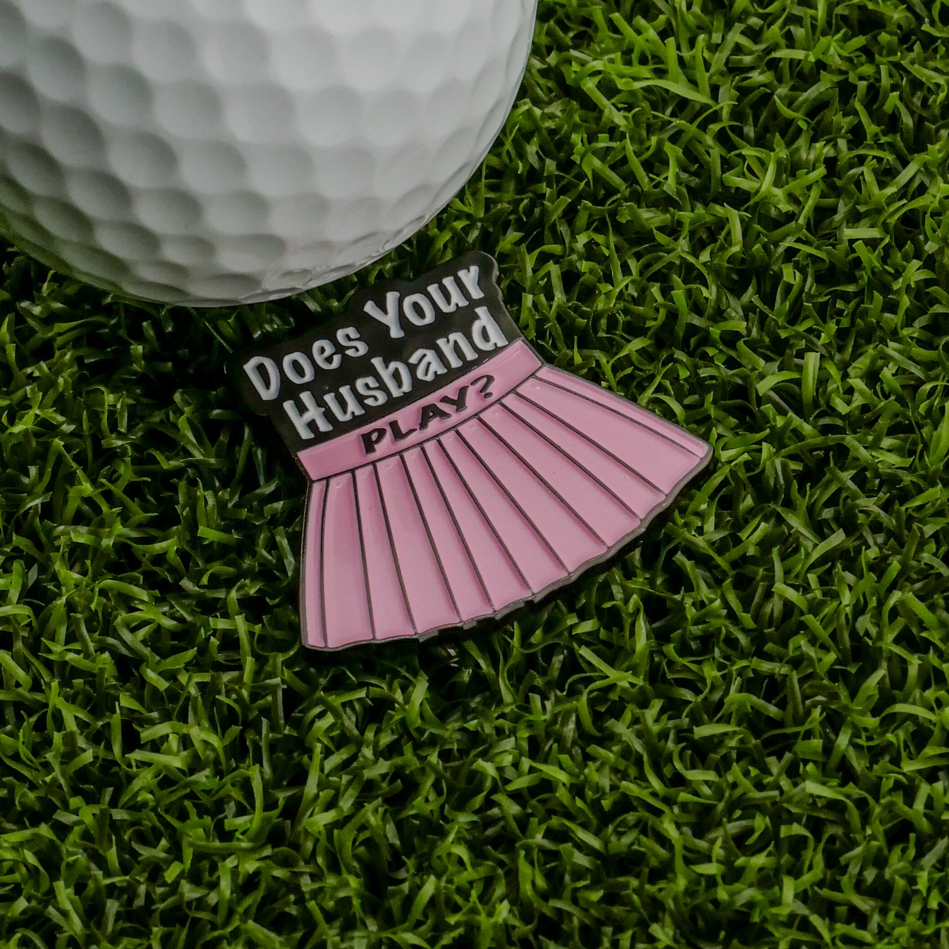 A golf ball rests on artificial grass beside Logie Joe's Golf "Does Your Husband Play?" Ball Marker—a pink, skirt-shaped marker that's a playful, funny golf gift for the course.