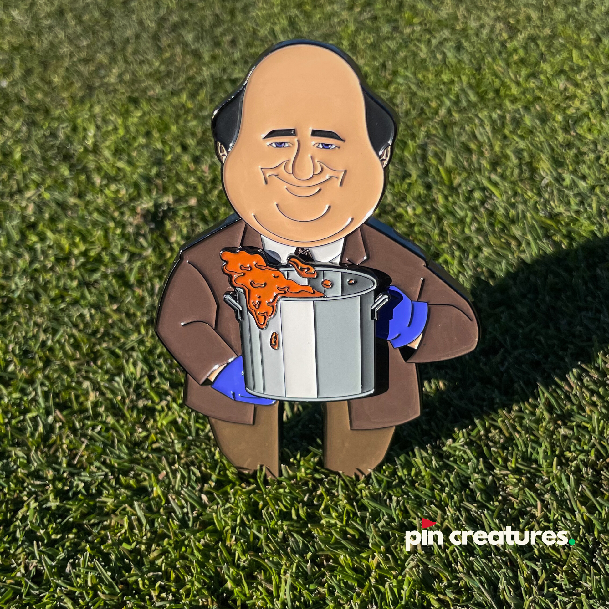 The Chili Cook Off Divot Tool by Pin Creatures features a cartoon bald man in a brown suit holding a pot of spilling orange chili, standing on grass. "Pin Creatures" appears in the bottom right—ideal for golf accessory fans.