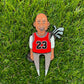 The GOAT 23 by Pin Creatures is a cartoon pin of a bald man in a red #23 basketball jersey holding golf clubs on green grass—an ideal golf accessory or unique ball marker for basketball and golf fans.
