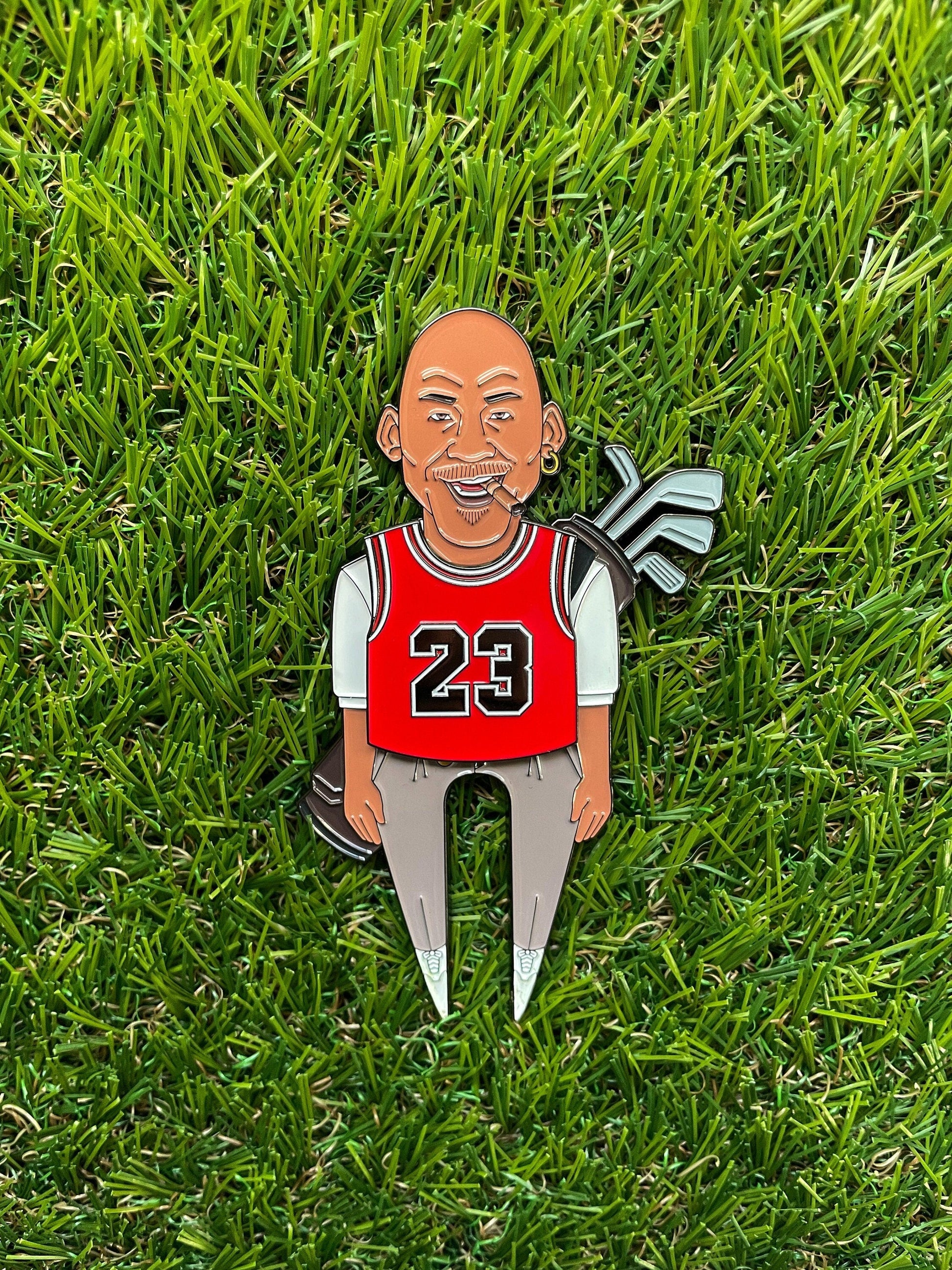 The GOAT 23 by Pin Creatures is a cartoon pin of a bald man in a red #23 basketball jersey holding golf clubs on green grass—an ideal golf accessory or unique ball marker for basketball and golf fans.