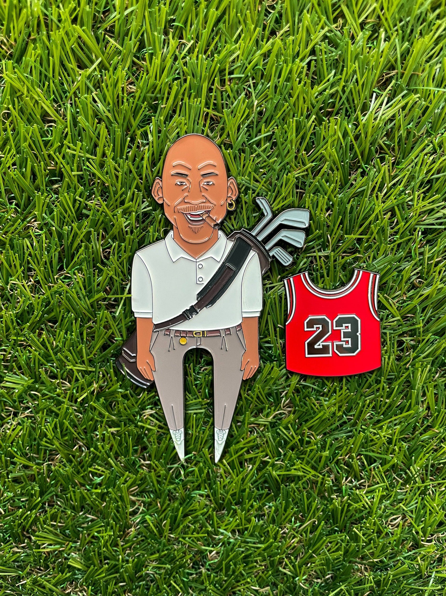 The GOAT 23 enamel pin by Pin Creatures features a smiling bald man in golf gear with clubs, standing on grass next to a red #23 jersey—an ideal golf accessory or unique ball marker for any fan.