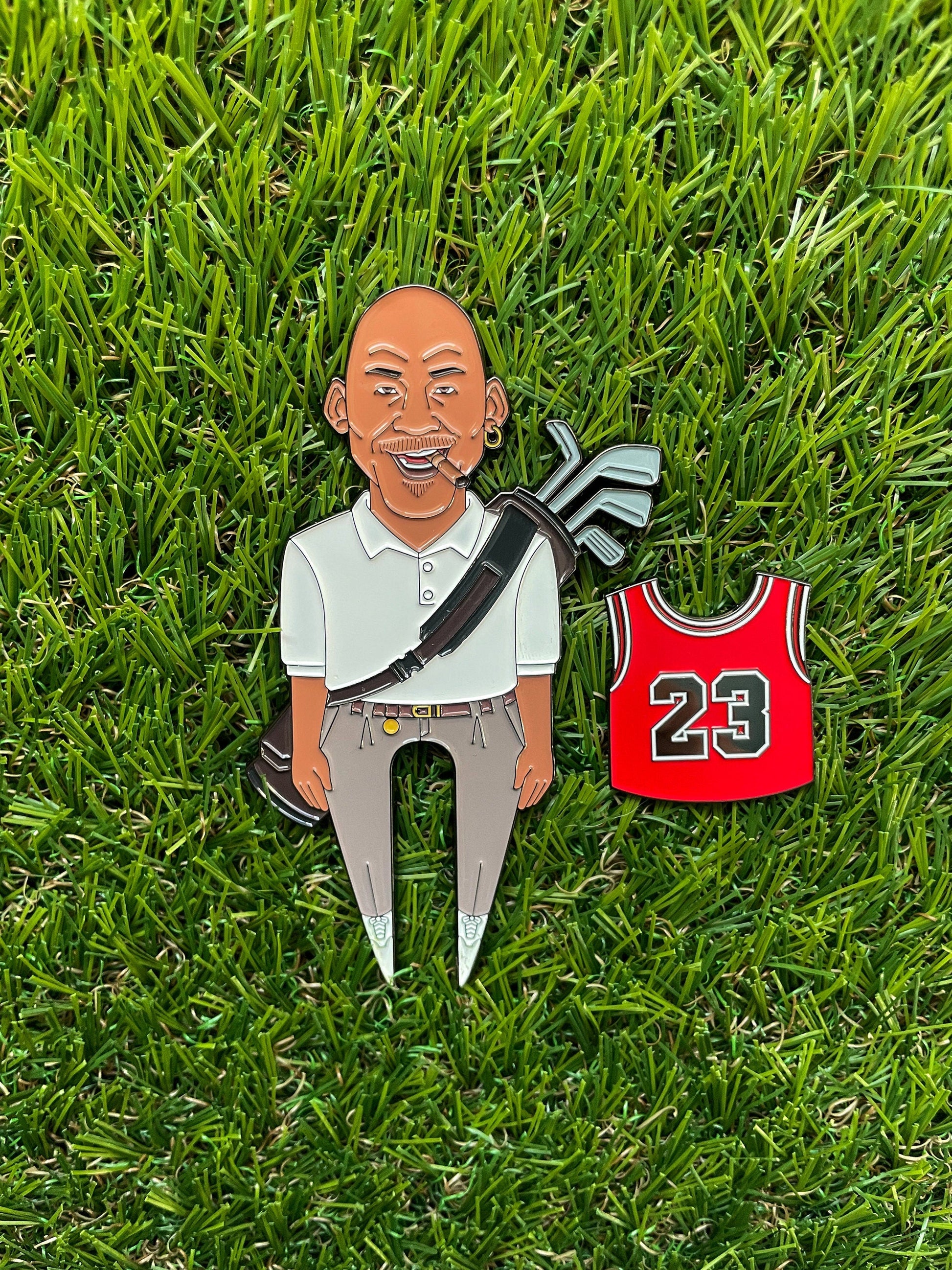 The GOAT 23 enamel pin by Pin Creatures features a smiling bald man in golf gear with clubs, standing on grass next to a red #23 jersey—an ideal golf accessory or unique ball marker for any fan.