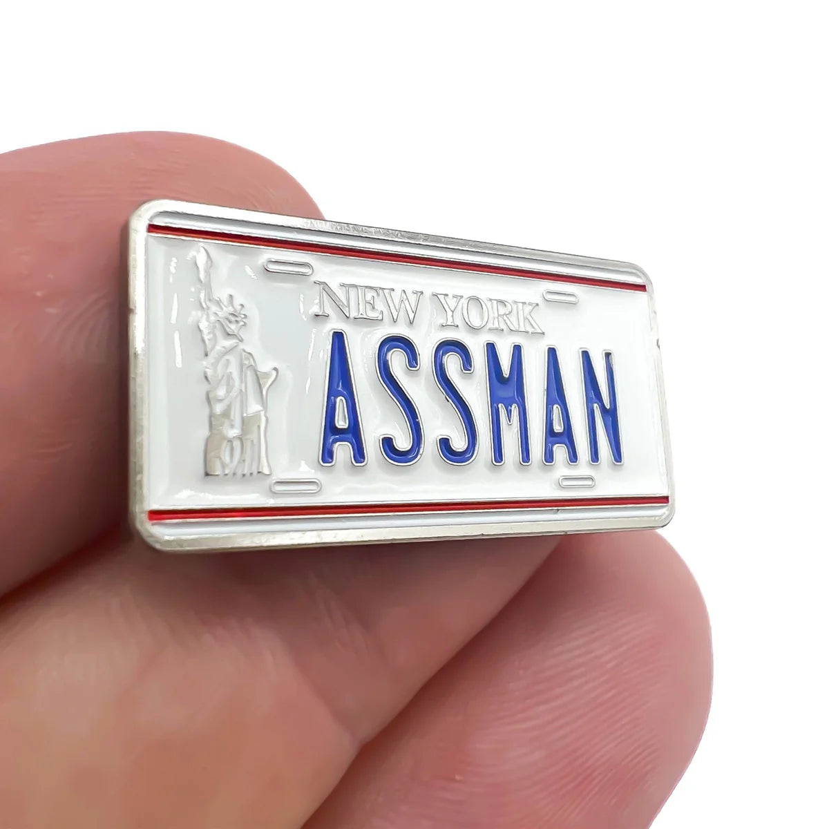A hand holds the Bogey Bros "Assman" novelty pin, shaped like a New York license plate with blue lettering and the Statue of Liberty—perfect for anyone who appreciates playful proctology humor.