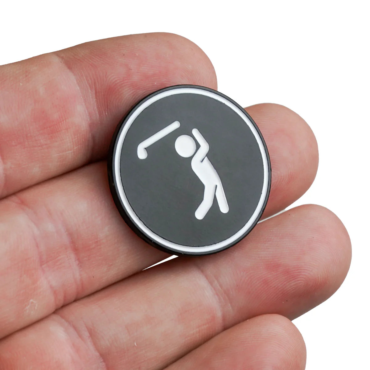 A close-up of a hand holding the Bogey Bros Bogeyman ballmarker, a round black and silver marker with a white stick figure golfer swinging a club.