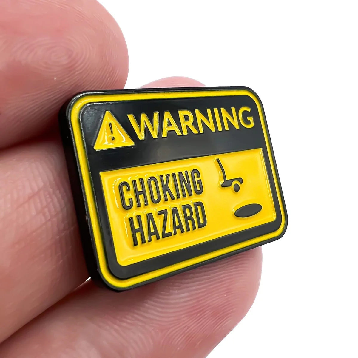 A hand holds Bogey Bros’ “Choking Hazard” pin, shaped like a yellow and black warning sign with a stick figure and small object—perfect for reminding golfers to avoid choking under pressure.