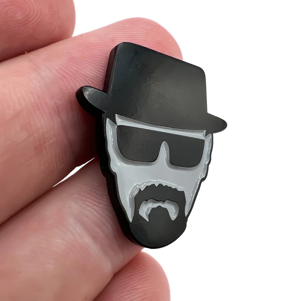 A hand holds the Bogey Bros "Heisenberg" pin, shaped like a stylized face with a black hat, sunglasses, beard, and mustache—capturing the iconic vibe—against a white background.