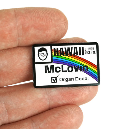 A hand holds a Bogey Bros McLovin novelty Hawaii driver license pin with a rainbow, cartoon face, name "McLovin," organ donor box, and a playful beer-themed nod perfect for fans.