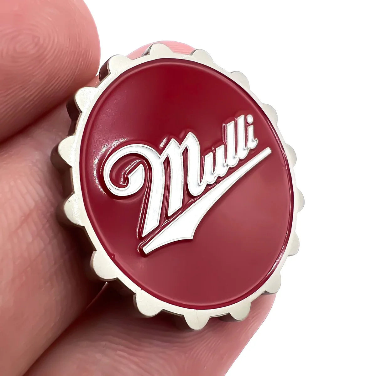 A hand holds a round, red, scalloped-edge pin resembling a bottle cap. The Bogey Bros Mulli pin features "Mulli" in white script—an ideal golf accessory for players ready to swing again.