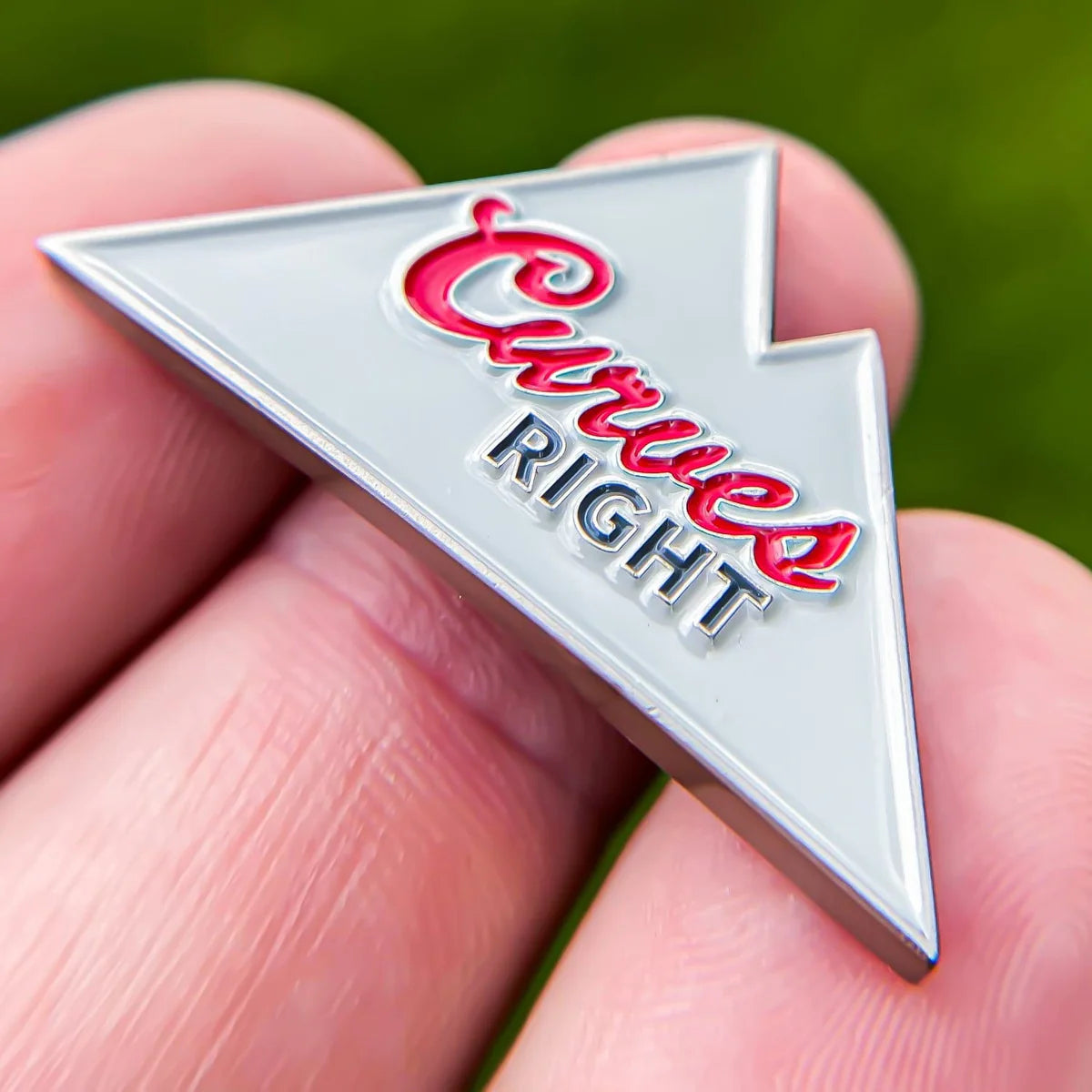 A close-up shows a hand holding the Bogey Bros "Curves Right" silver enamel pin, shaped like a right-pointing arrow with "Curves RIGHT" in red and white—ideal for golfers familiar with a slice. Blurred grass is visible in the background.