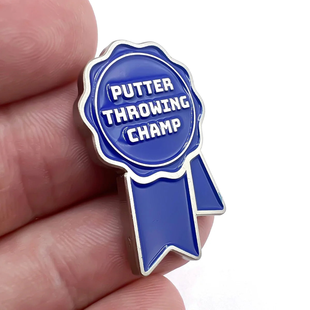 A hand holds the Bogey Bros "Putter Throwing Champ" blue enamel pin—shaped like an award ribbon with white text—making it a perfect golf gift for anyone who loves a good golf joke.