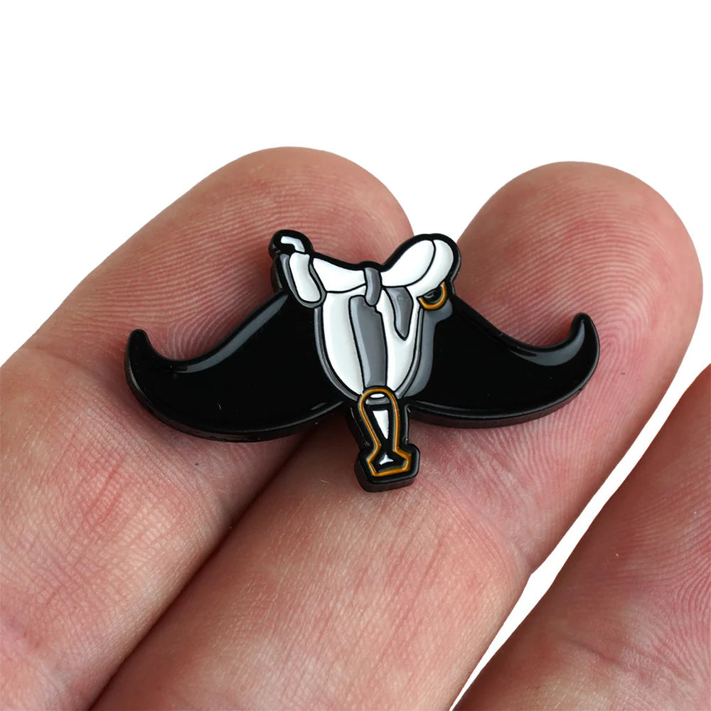 A hand holds the "Fore!" enamel pin by Bogey Bros, shaped like a black mustache with a white and gold saddle—an ideal accessory for fans of golf humor and quirky golfing style.
