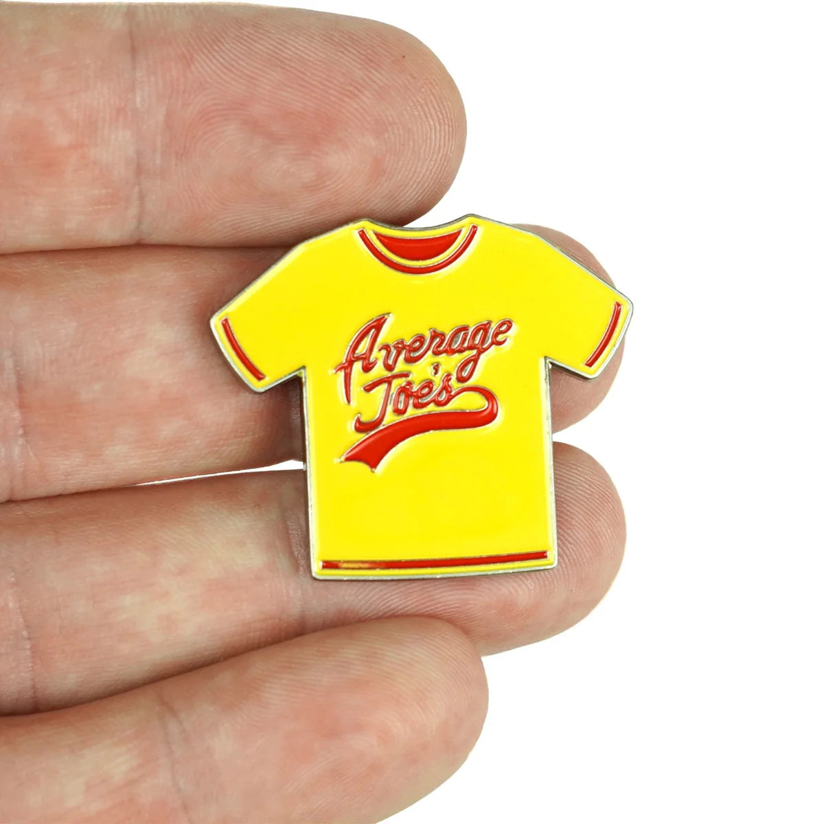 A hand holds a Bogey Bros "Average Joes" enamel pin shaped like a yellow T-shirt with red accents—ideal for any dodgeball fan who loves the classic motto, “If you can dodge a wrench, you can dodge a ball.”.