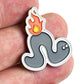 A hand holds the Bogey Bros "Worm Burner" pin, featuring a cartoon wave with fire and yellow flame—blending water and fire in a fun design that's a unique golf gift for players who love golf humor.