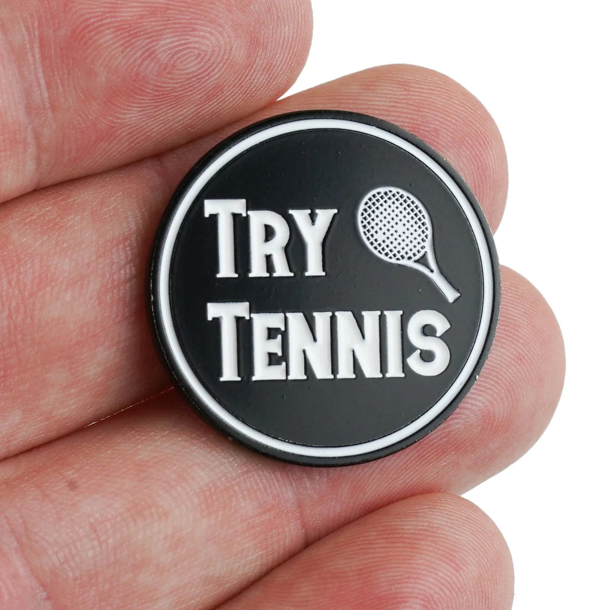A hand holds a round black "Try Tennis" pin by Bogey Bros, featuring white text and a small tennis racket graphic—ideal for anyone interested in starting the sport.
