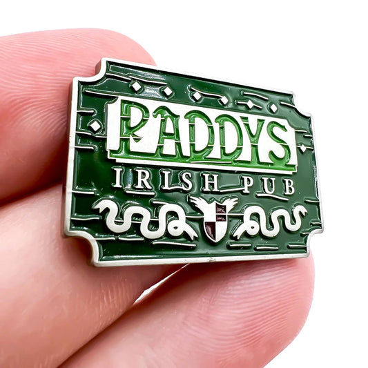 A hand holds the Paddy's Pub green enamel pin by Bogey Bros, featuring decorative lines, two white snakes, and a small shield—an eye-catching golf accessory that doubles as a stylish ball marker.