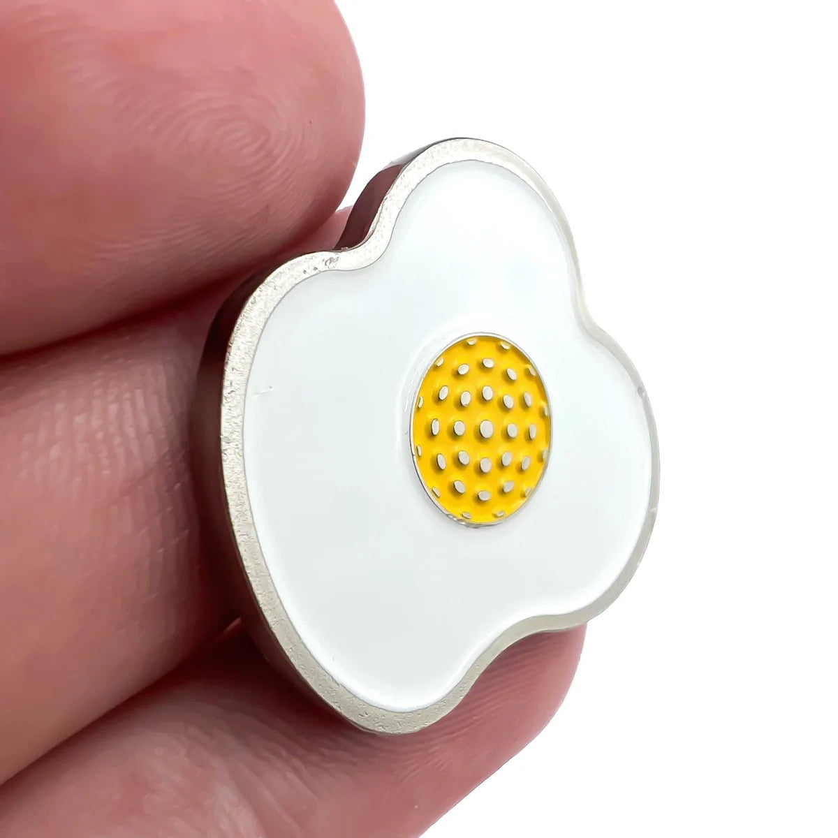 A hand holds the Bogey Bros Fried Egg pin, featuring a quirky fried egg design with a white outer area and a yellow, textured yolk—perfect for adding playful style to any look.