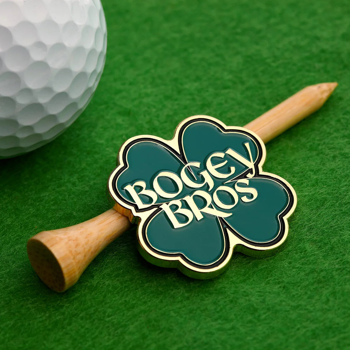 The Gettin' Lucky pin by Bogey Bros, shaped like a clover, sits on a wooden golf tee beside a white golf ball on green turf.