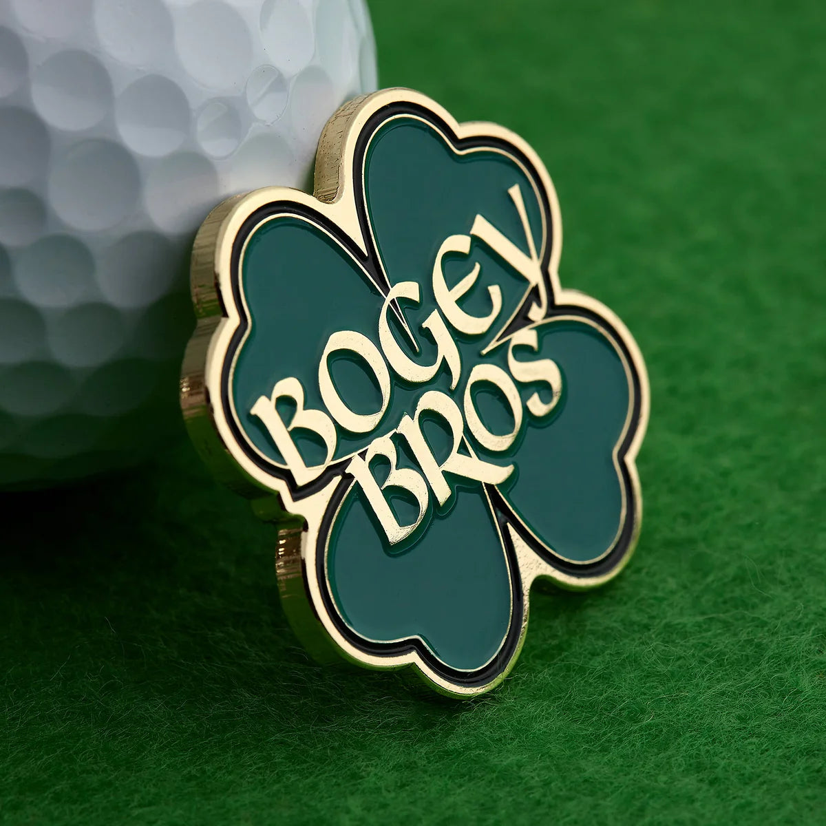 The Bogey Bros "Gettin' Lucky" golf ball marker, shaped like a gold and green four-leaf clover, rests on green turf next to a white golf ball.