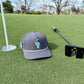 A gray Logie Joe's Golf Astronaut Walker Golf Hat with a cartoon astronaut and built-in magnetic ball marker sits by a black putter near the hole with a white flag, set against trees and blue sky.