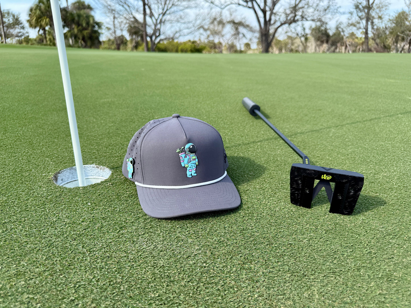 A gray Logie Joe's Golf Astronaut Walker Golf Hat with a cartoon astronaut and built-in magnetic ball marker sits by a black putter near the hole with a white flag, set against trees and blue sky.