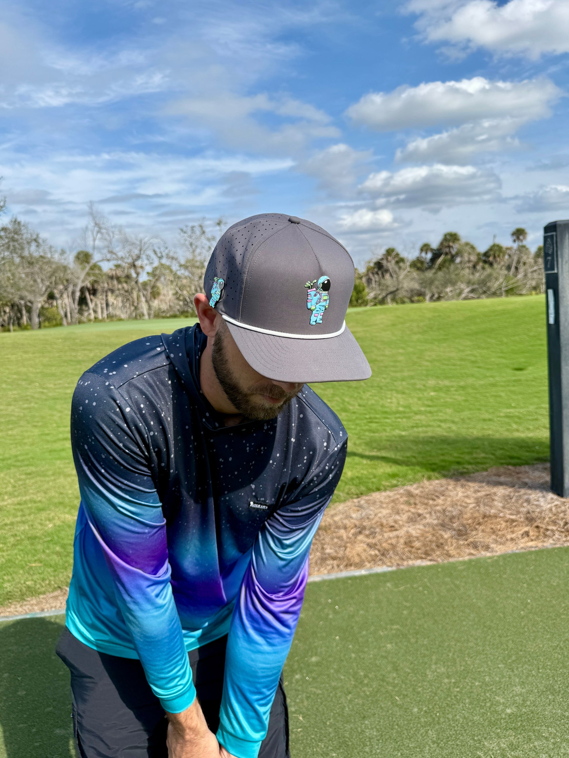 A man in a Logie Joe's Golf Astronaut Walker Golf Hat—Snapback Cap with built-in magnetic ball marker holder—is preparing to hit a golf ball on a green course under a partly cloudy sky.