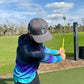 Wearing the gray Astronaut Walker Golf Hat by Logie Joe's Golf, featuring a built-in magnetic ball marker holder, a golfer swings on a grassy course beneath partly cloudy skies, with trees and green landscape in the background.